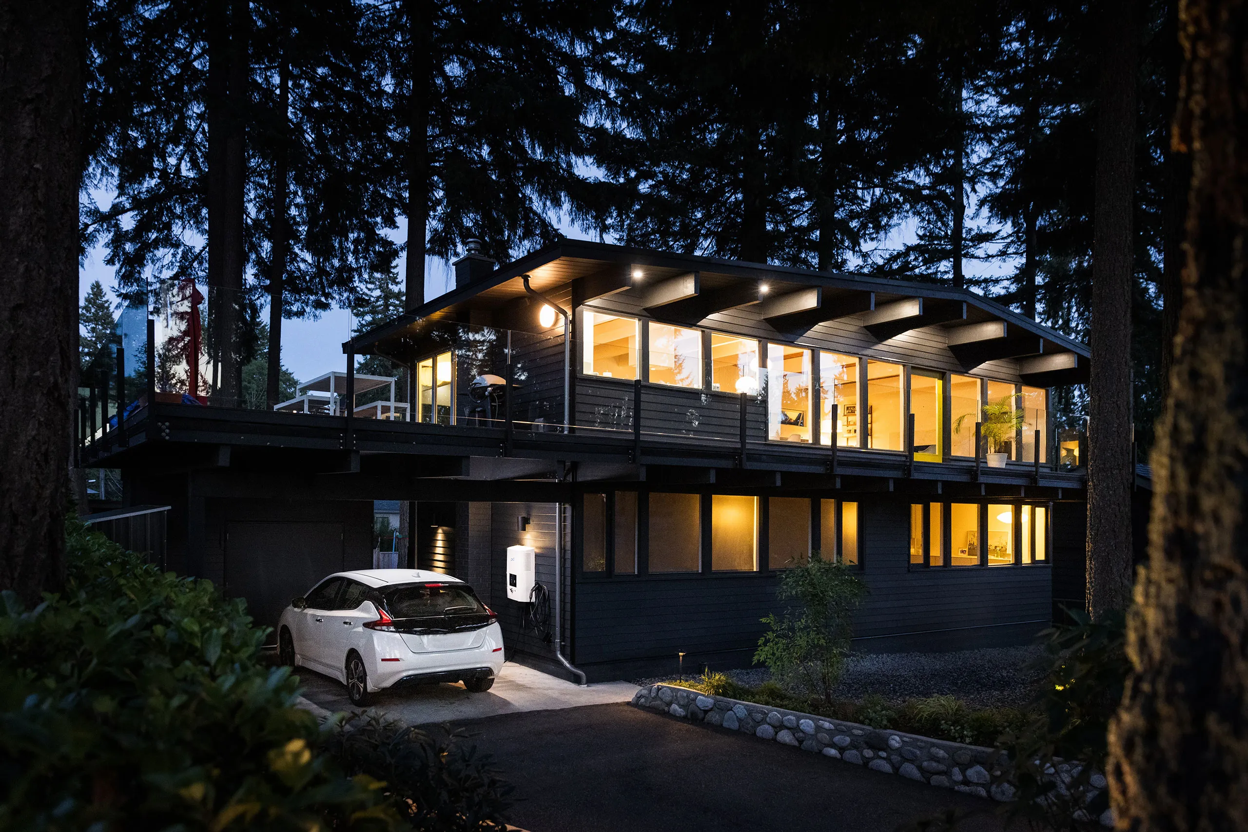 Stimmungsvolles energetisch modernes Haus in der Abenddämmerung in einem Waldstück mit E-Auto in der Einfahrt